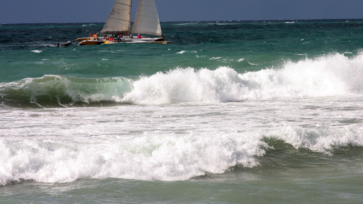 A trimaran sails across strong wind beyond breaking coastal waves