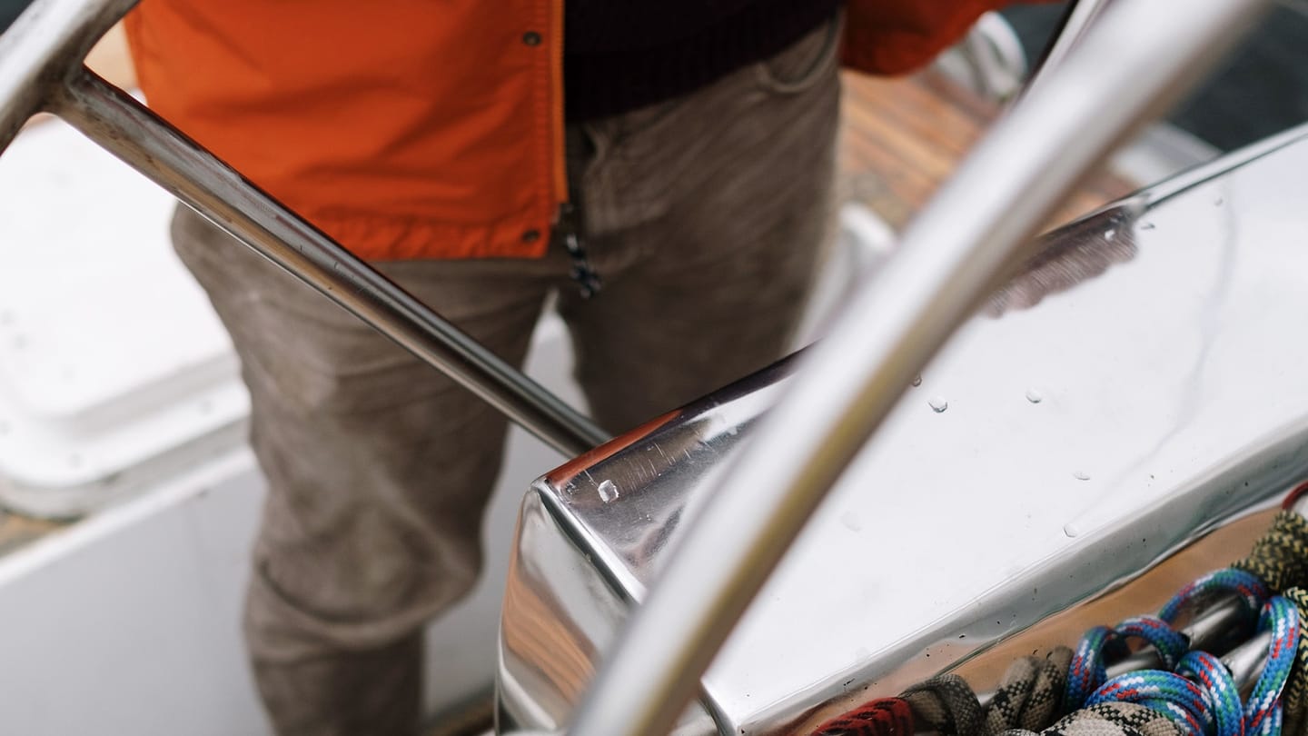 A sailor grips a yacht wheel with deck lines visible behind it.