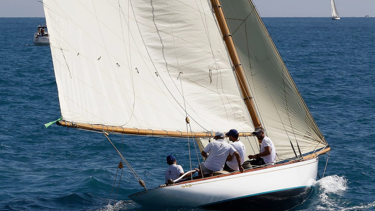 Three sailors sit in a yacht cockpit under a bright sky.