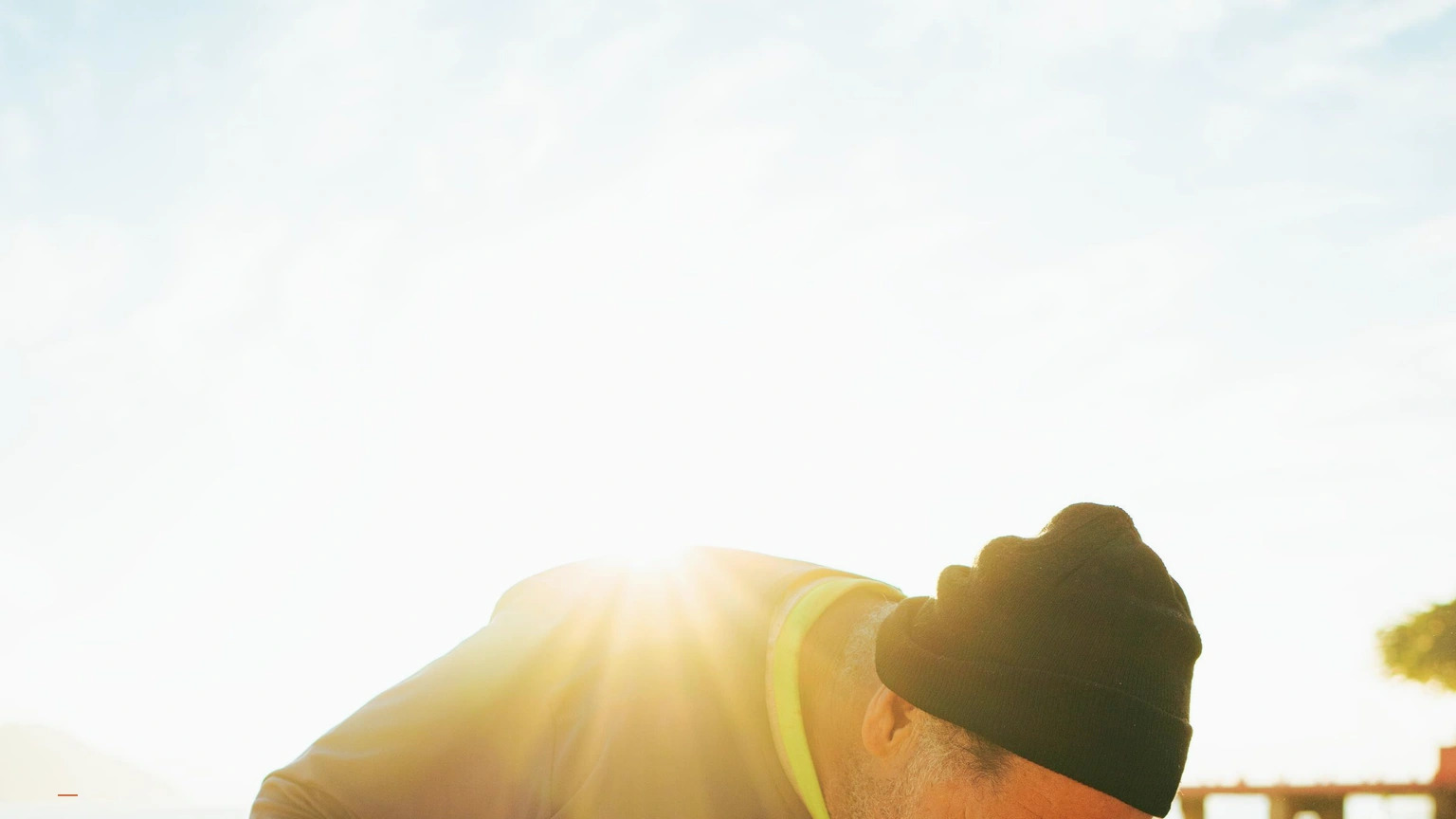 Person in a beanie leaning forward in strong backlit sunlight while working near the water