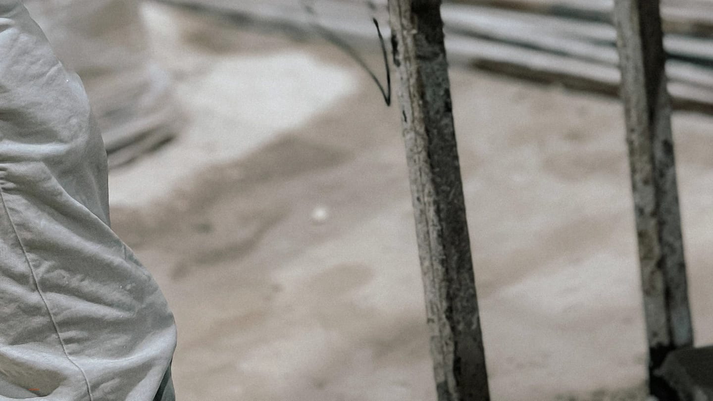 Blurred boatyard scene showing a person in coveralls beside vertical metal stands supporting a hull