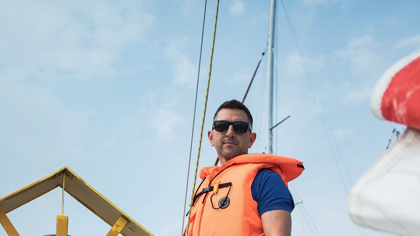A sailor in an orange lifejacket stands beside the rigging of a sailboat.
