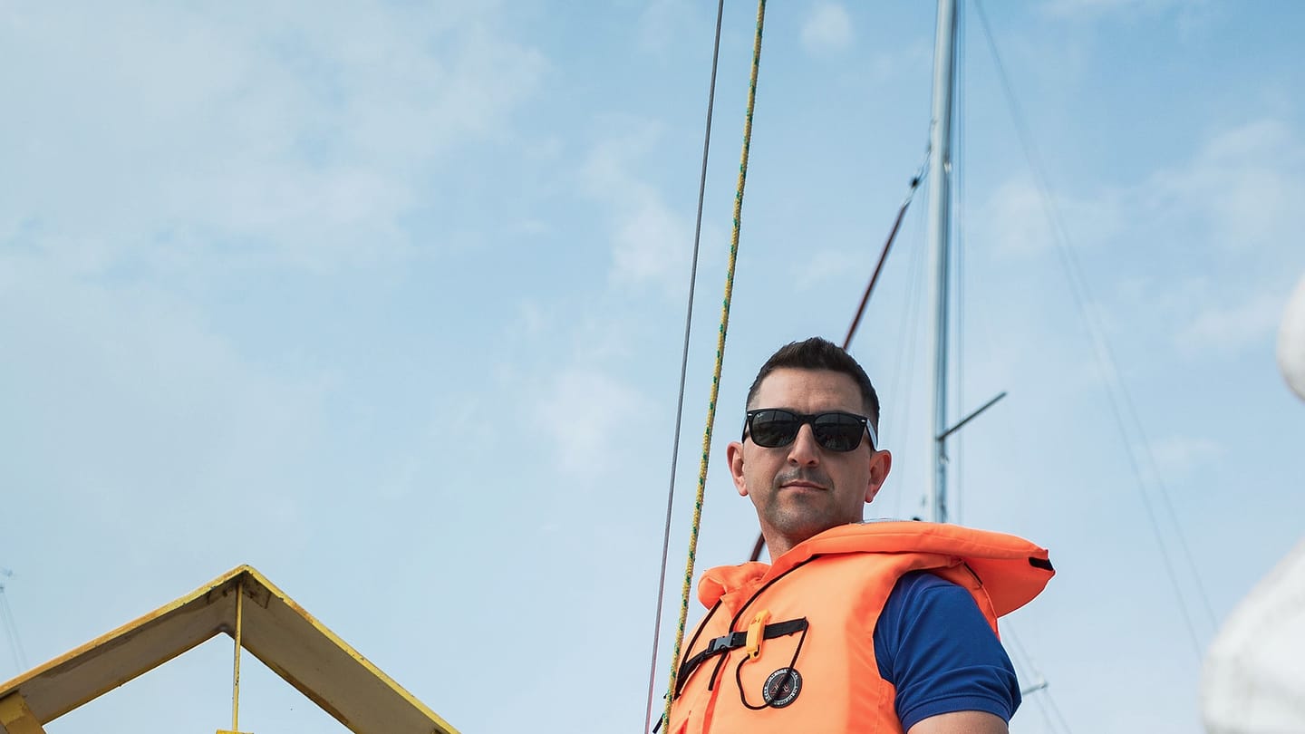 A sailor in an orange lifejacket stands beside rigging in a marina.