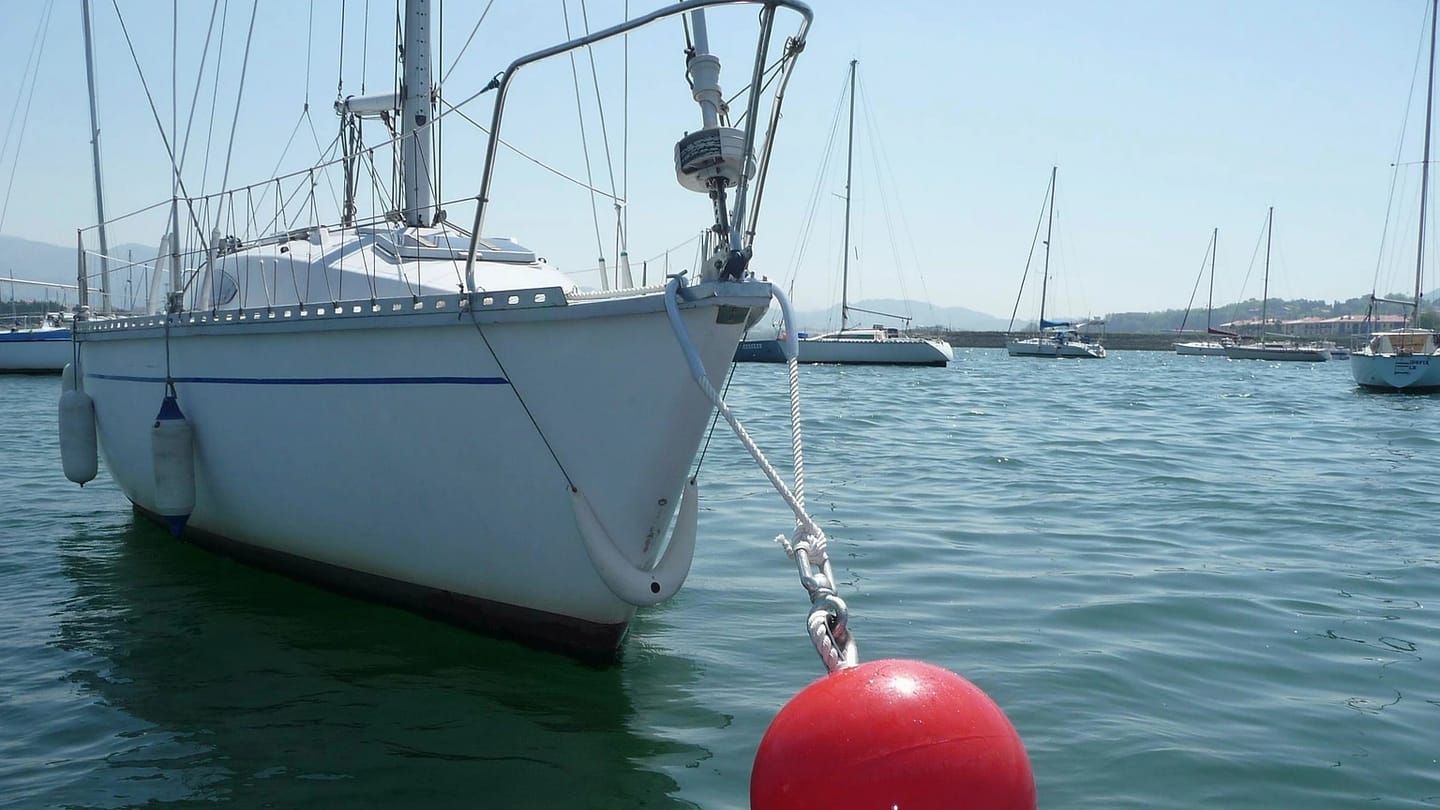 A sailboat is tied to a bright red mooring buoy in calm water.