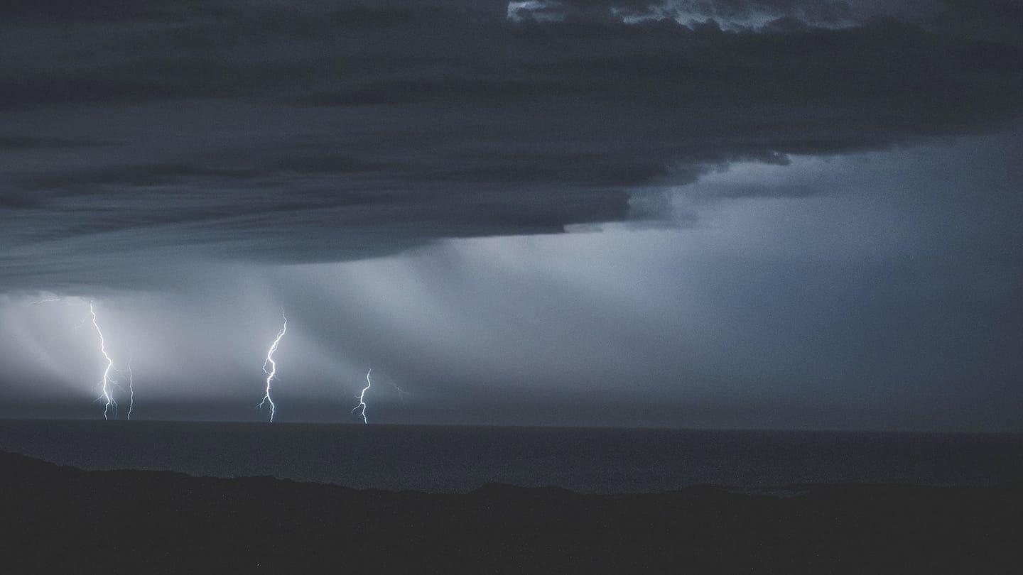 Lightning descends from dark storm clouds over open water.