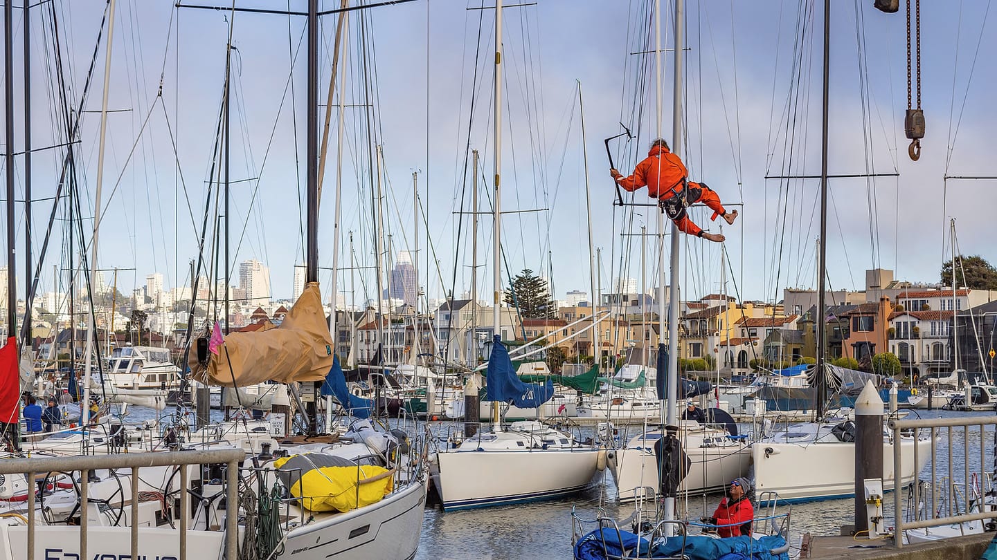 A marine technician in an orange harness works aloft among marina masts.