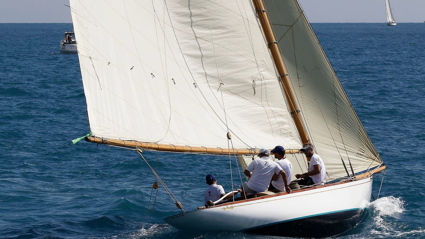 Three sailors sit in the cockpit of a white yacht underway.