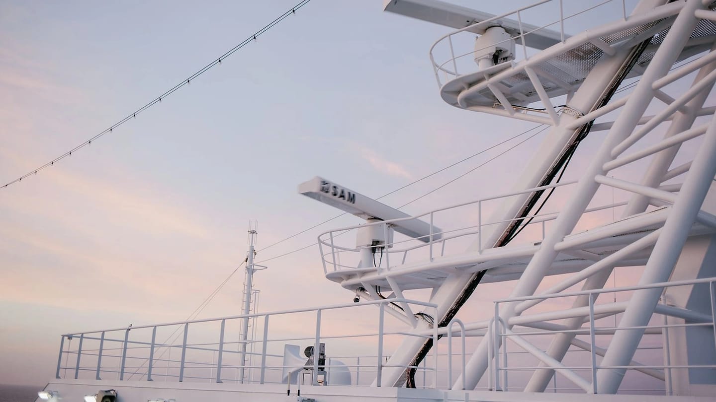 A large vessel's radar antenna and deck equipment are shown against a pastel sky.