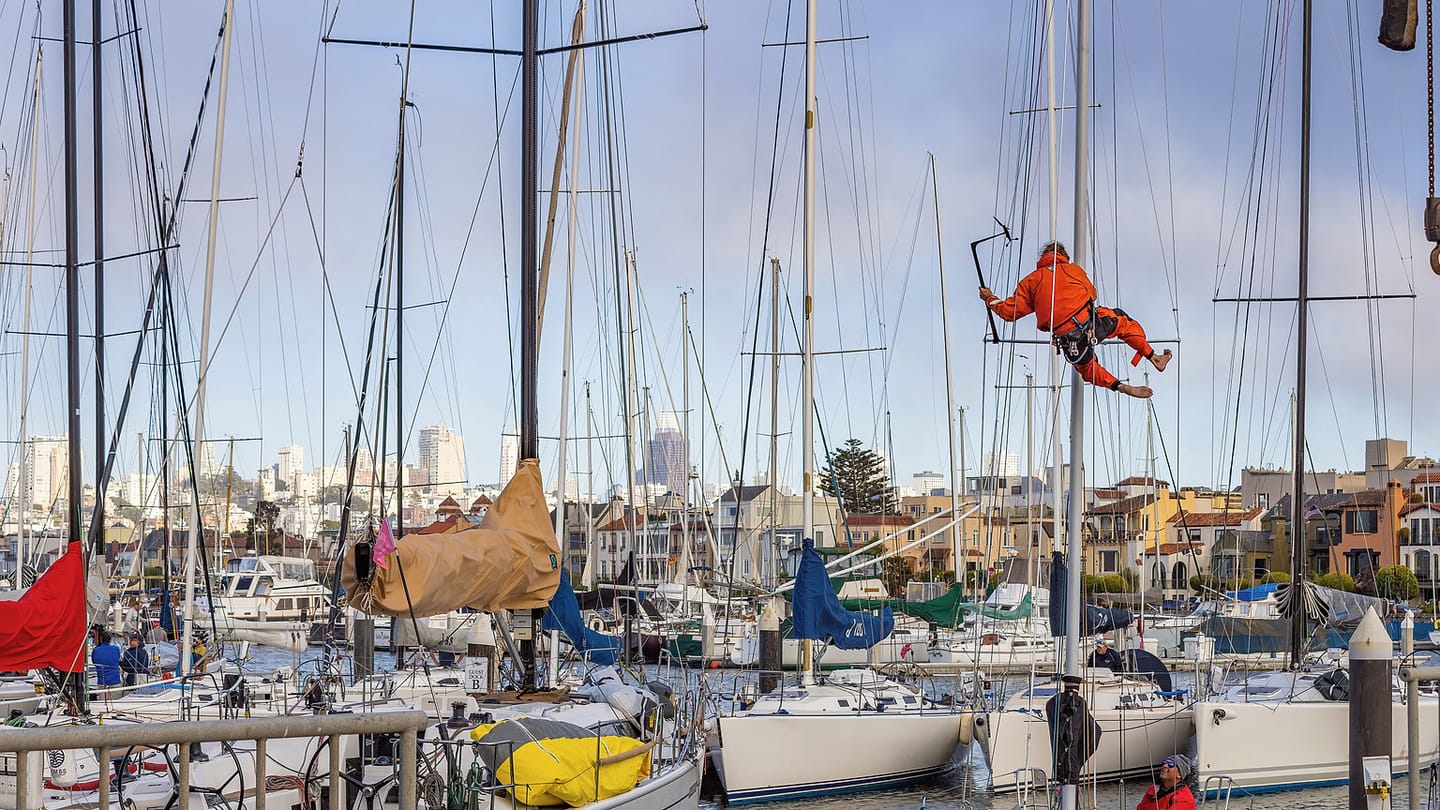 A marine technician in an orange harness works aloft beside a mast.