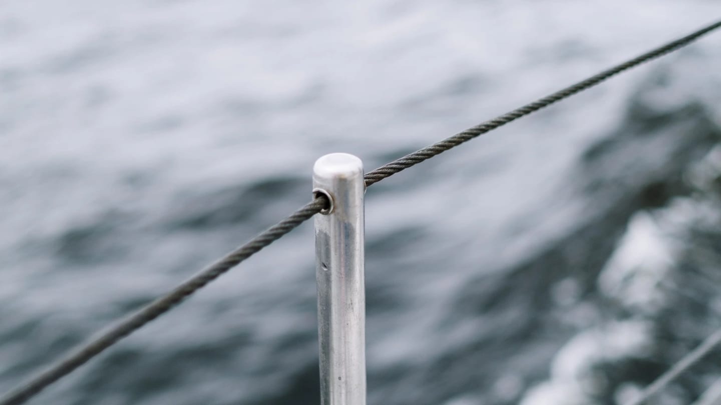 Two bare stainless lifelines passing through a vertical stanchion beside a sailboat deck, with open water beyond.