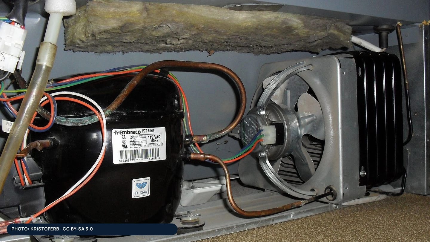 A refrigerator compressor, condenser coil, fan, and copper tubing behind an access panel