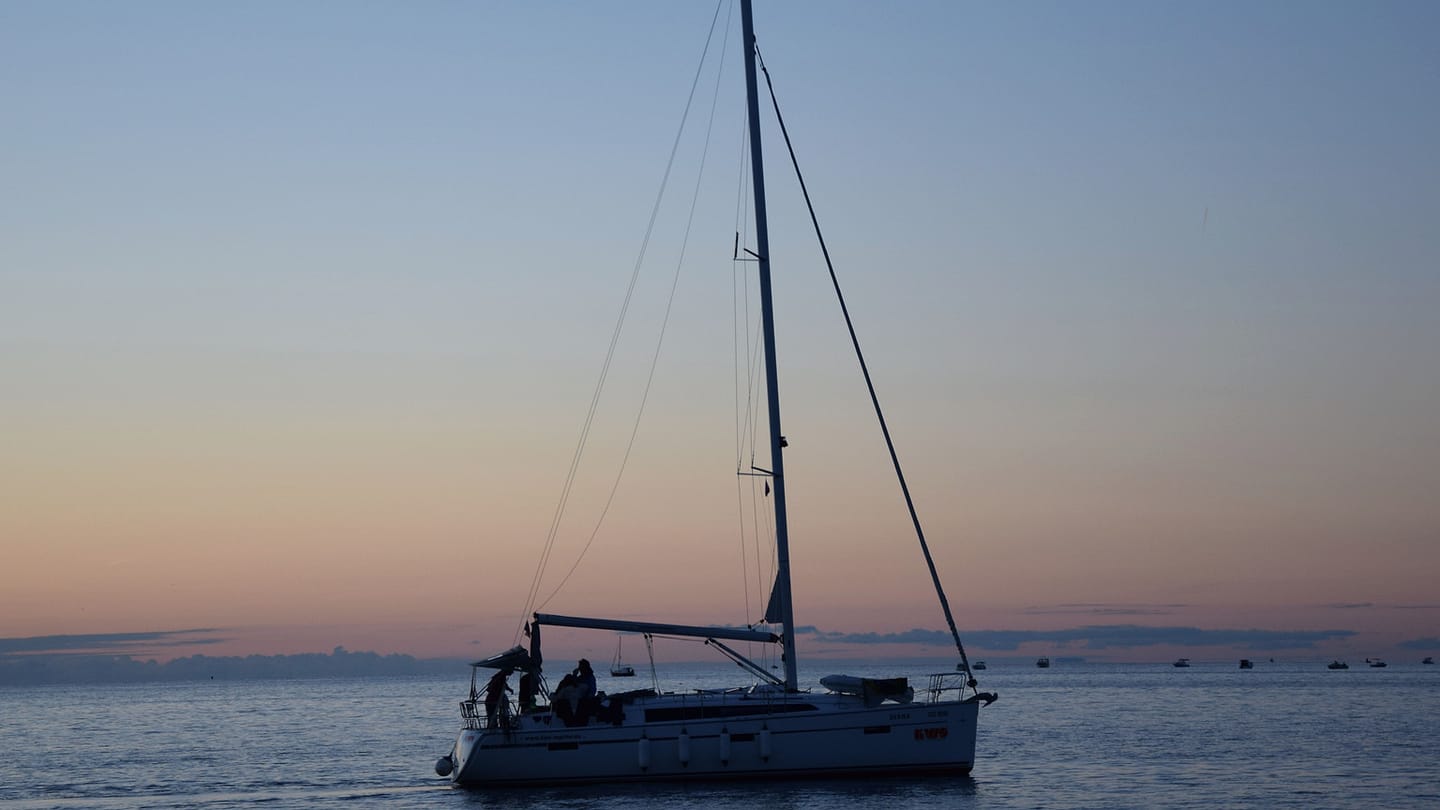 A sailing yacht moves across calm water after sunset.