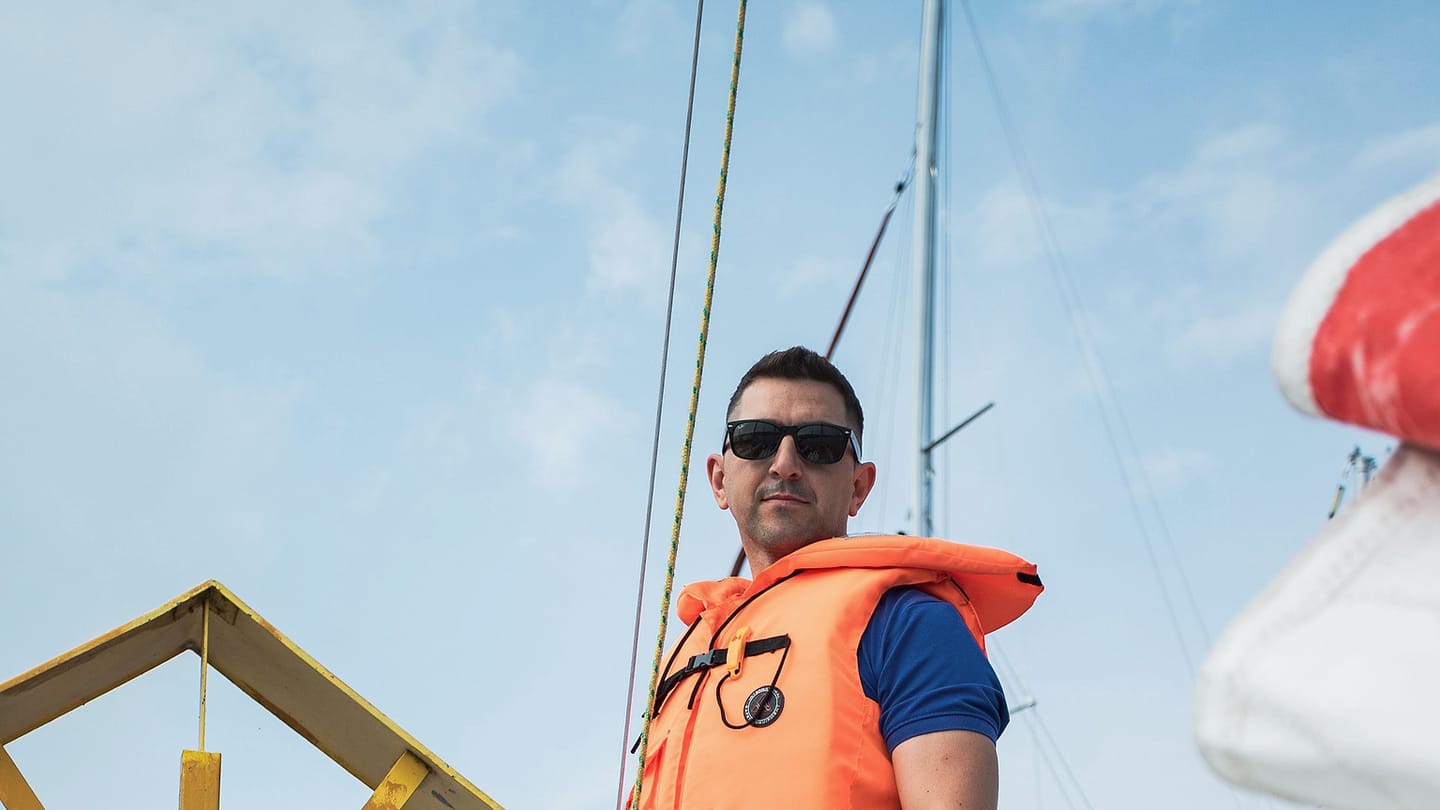 A sailor in an orange lifejacket stands on a sailboat with open sky behind him.