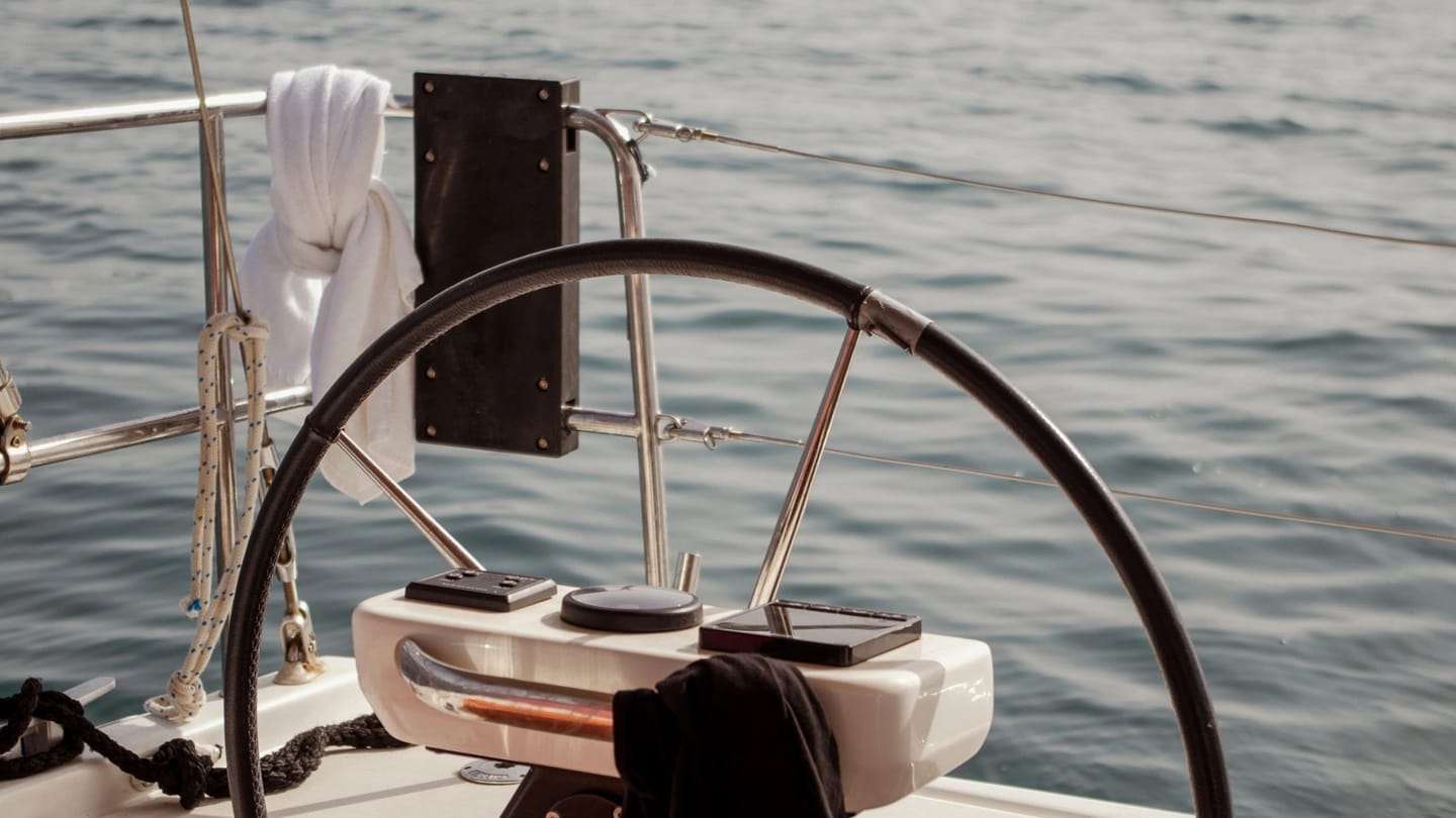 Black steering wheel and white pedestal in the cockpit of a modern sailboat, with open water behind the stern.