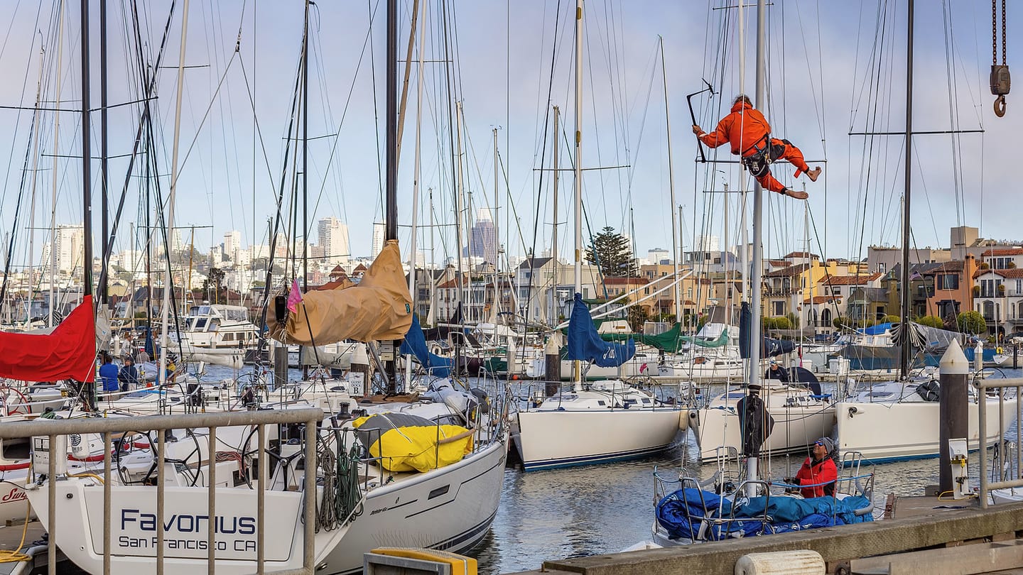 A marine technician in an orange harness works high beside a sailboat mast.