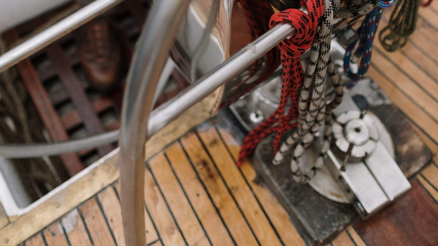 A sailor in an orange jacket holds a large wheel in a yacht cockpit.