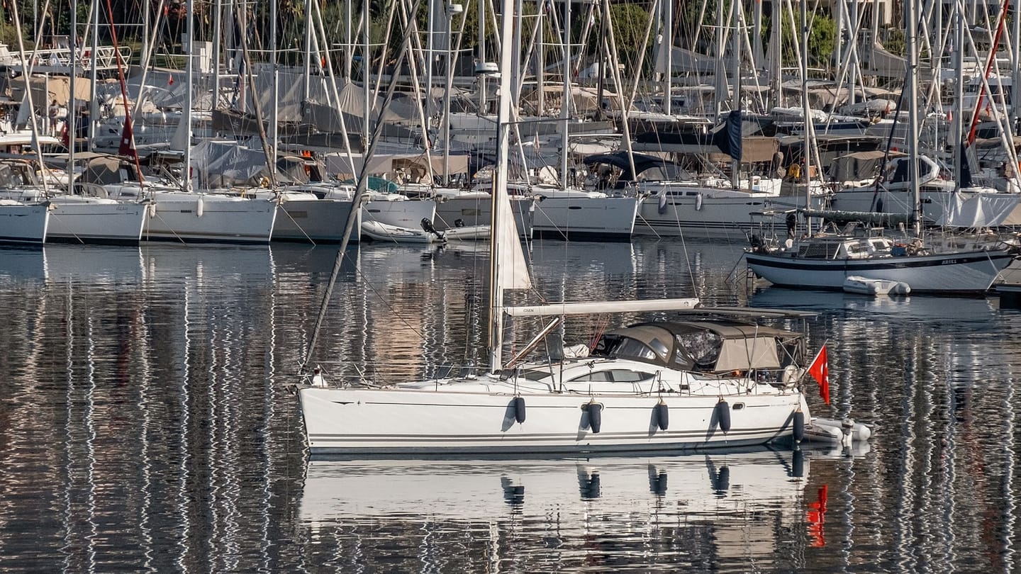 A white sailboat moves slowly out of a marina lined with yachts.
